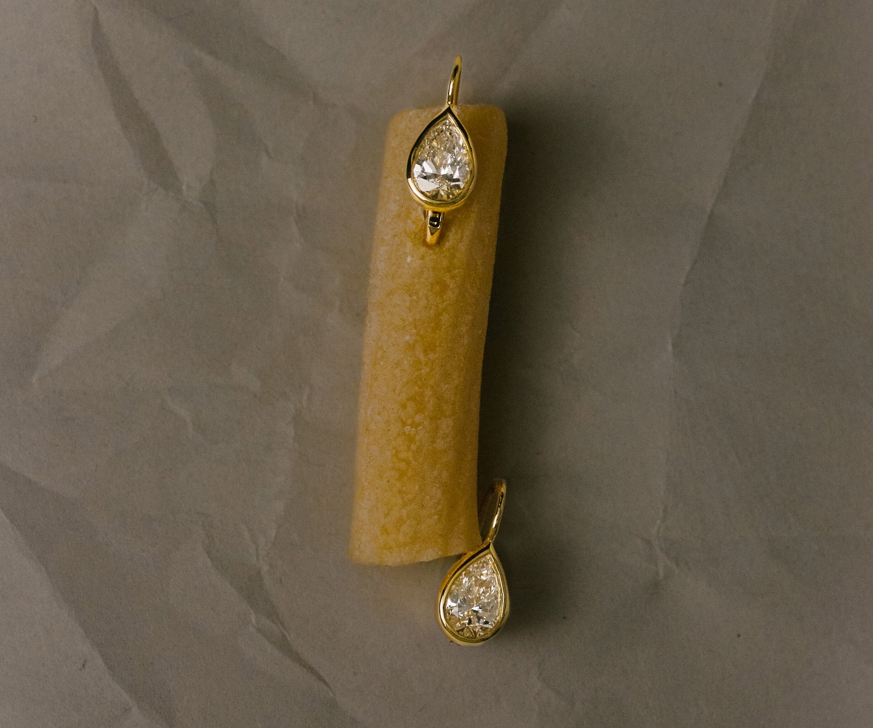 A pair of Kinn 14k gold Harper Pear Diamond Drop Earrings with one hooked onto a piece of penne pasta against a crinkled gray background