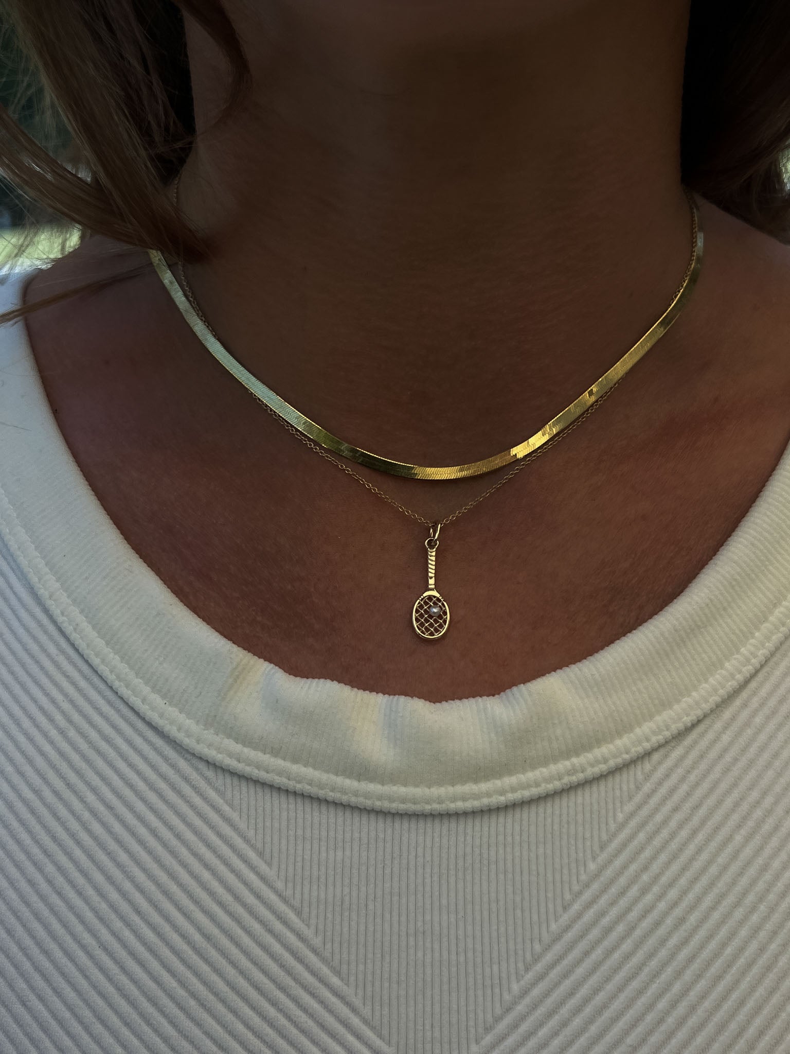 A Kinn x Sunset Tennis Club 14k Gold Pearl Ball And Racquet Charm Necklace layered with a Carter Herringbone Chain Necklace