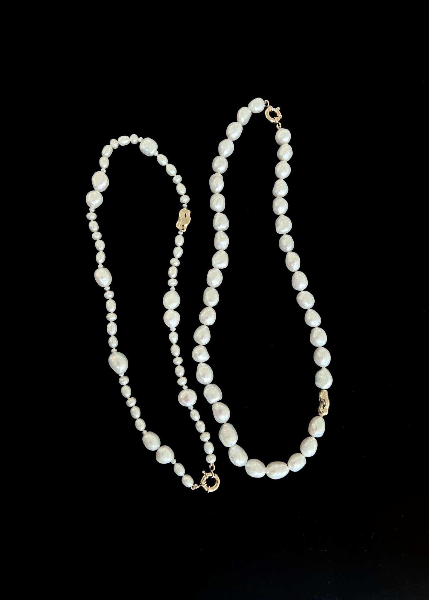 A Kinn Studio 14k Gold Lucy Baroque Pearl Necklace and Ruth Baroque Pearl Necklace laid next to each other against a black background