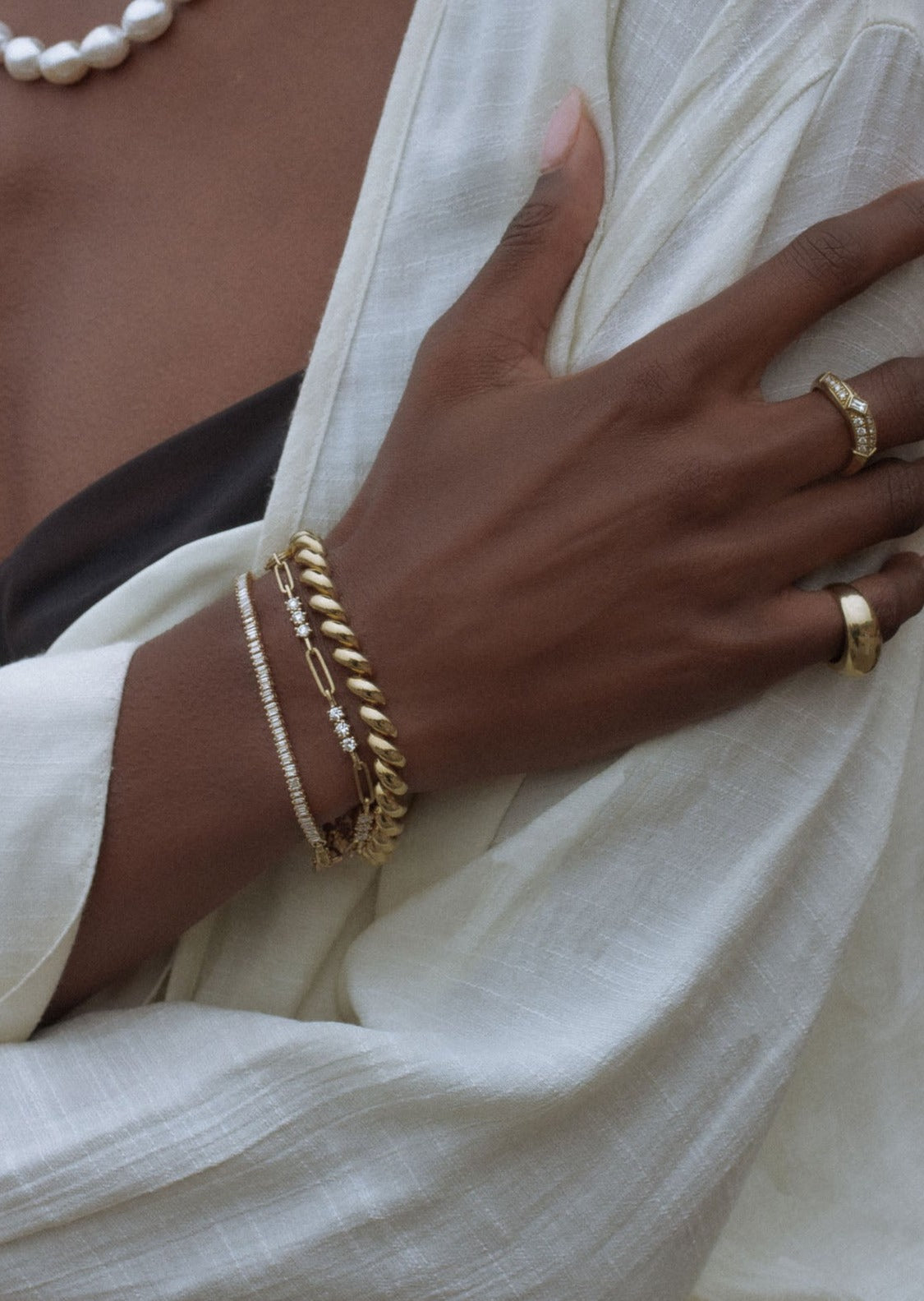 woman in a white top wearing a Kinn Studio 14k Gold Hampshire House Bracelet layered with a Karina Trio Bracelet Diamond, and a Serena Baguette Diamond Bracelet