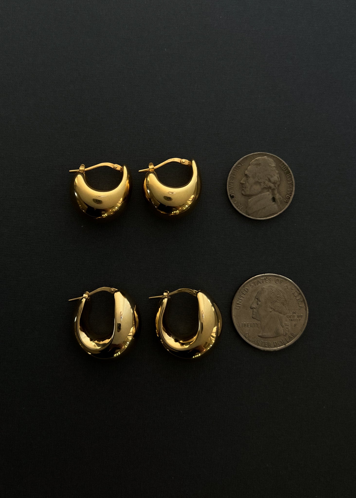Kinn 14k Gold Bold Teardrop Hoop Earrings and 14k Gold Petite Bold Teardrop Hoops laying flat with a nickel and quarter against a black background to show size comparison