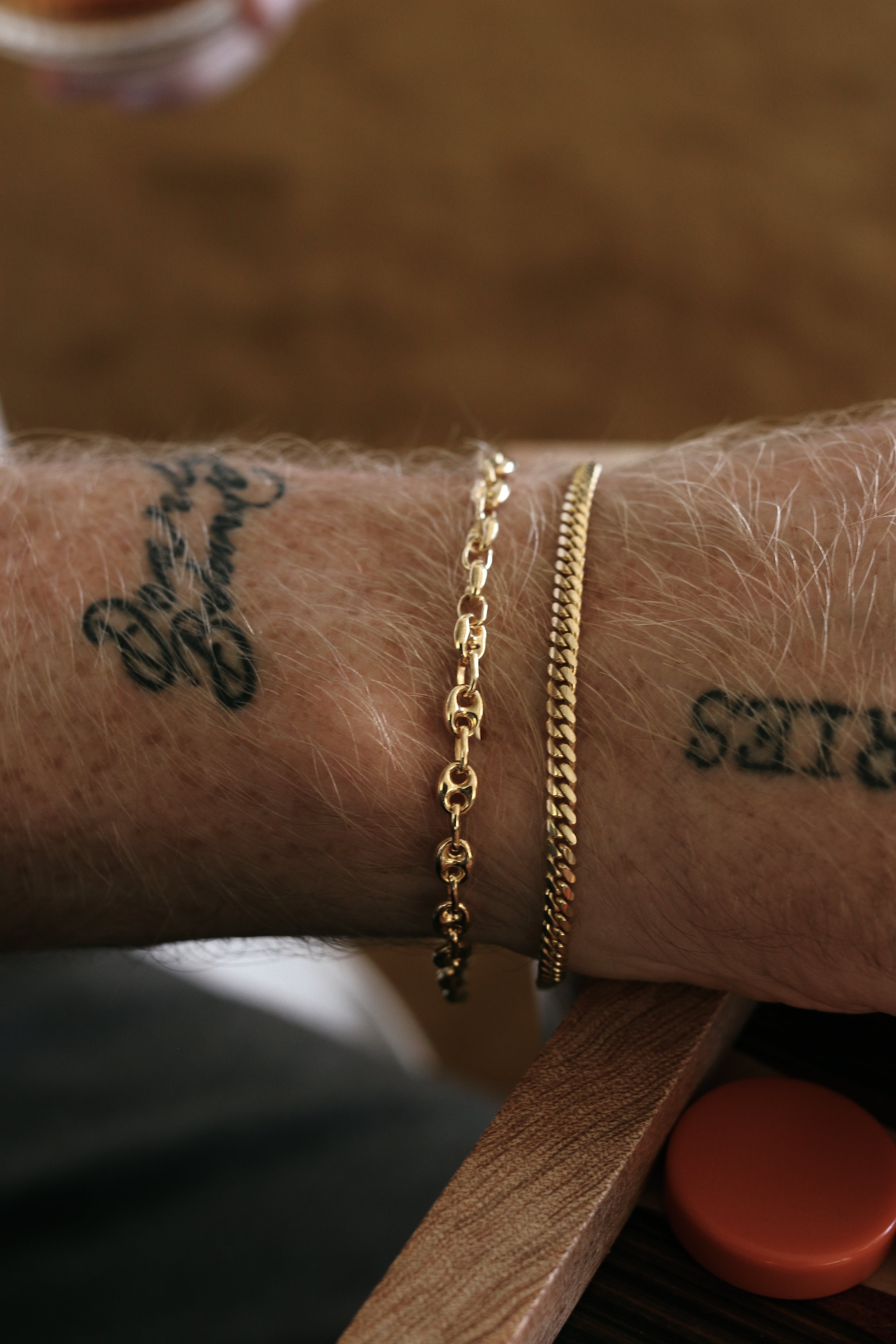 A close up of a man's arm wearing a Kinn 14k Gold Men's Petite Puffed Mariner Chain Bracelet stacked with a Familia Cuban Chain Bracelet