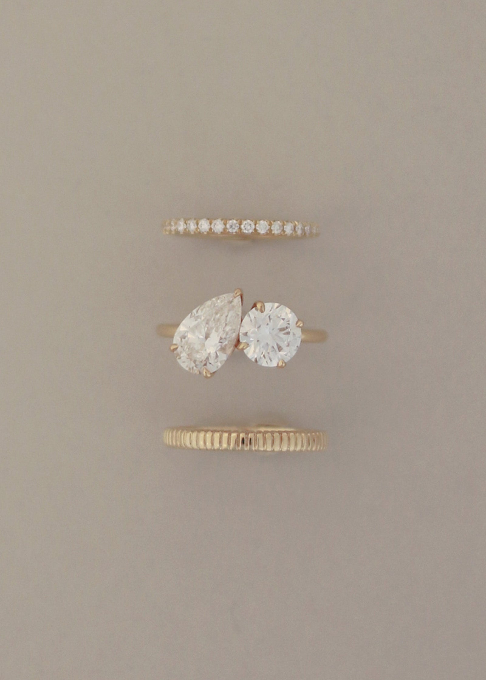 A Kinn 14k gold Toi et Moi Annette Engagement Ring featuring a pear shaped diamond and a round diamond shown with  a Dahlia Pavé Diamond Band and Annabelle Band stacked upright against a gray background