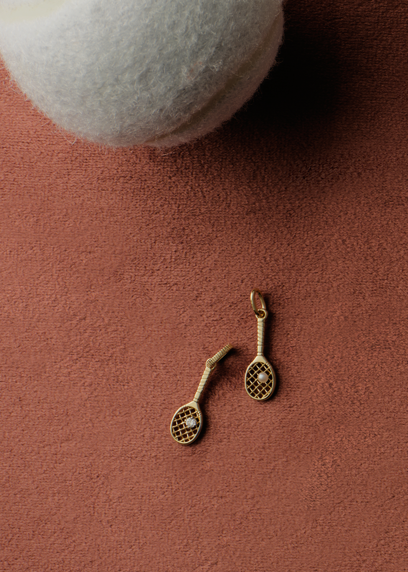 Two Kinn x Sunset Tennis Club 14k Gold Pearl Ball And Racquet Charms laying on a clay background next to a white tennis ball