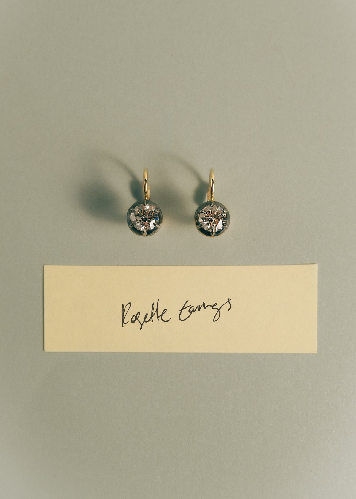 A pair of Kinn 14k Gold Rosette Black Rhodium Round Diamond Drop Earrings on a beige paper background with a piece of paper labeled "Rosette Earrings" 
