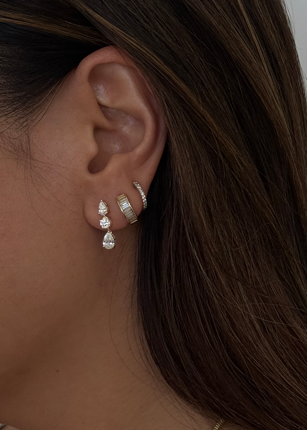 Close up of a woman's ear wearing a Kinn 14k Gold Third Time's a Charm Drop Earrings Pear 2.7ctw Diamond layered with a Bobbi Baguette Diamond Huggie and a Pave Huggie