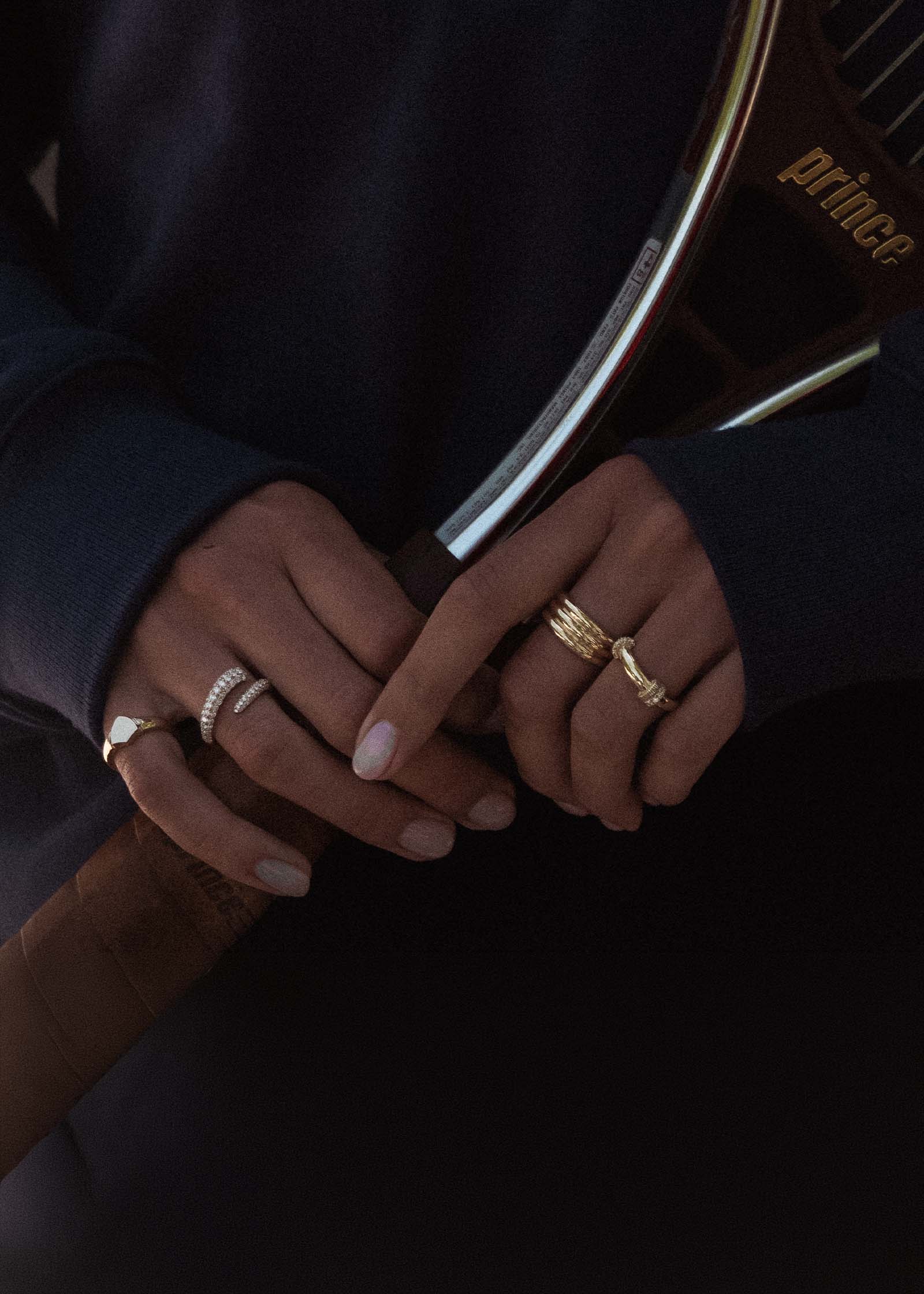 woman in a navy sweatshirt and holding a Prince tennis racquet wearing a Kinn x Prince 14k Gold Rally Pavé Diamond Ring, Francoise Ellipse Ring, Pave Wrap Around Ring, and Ace Shield signet ring