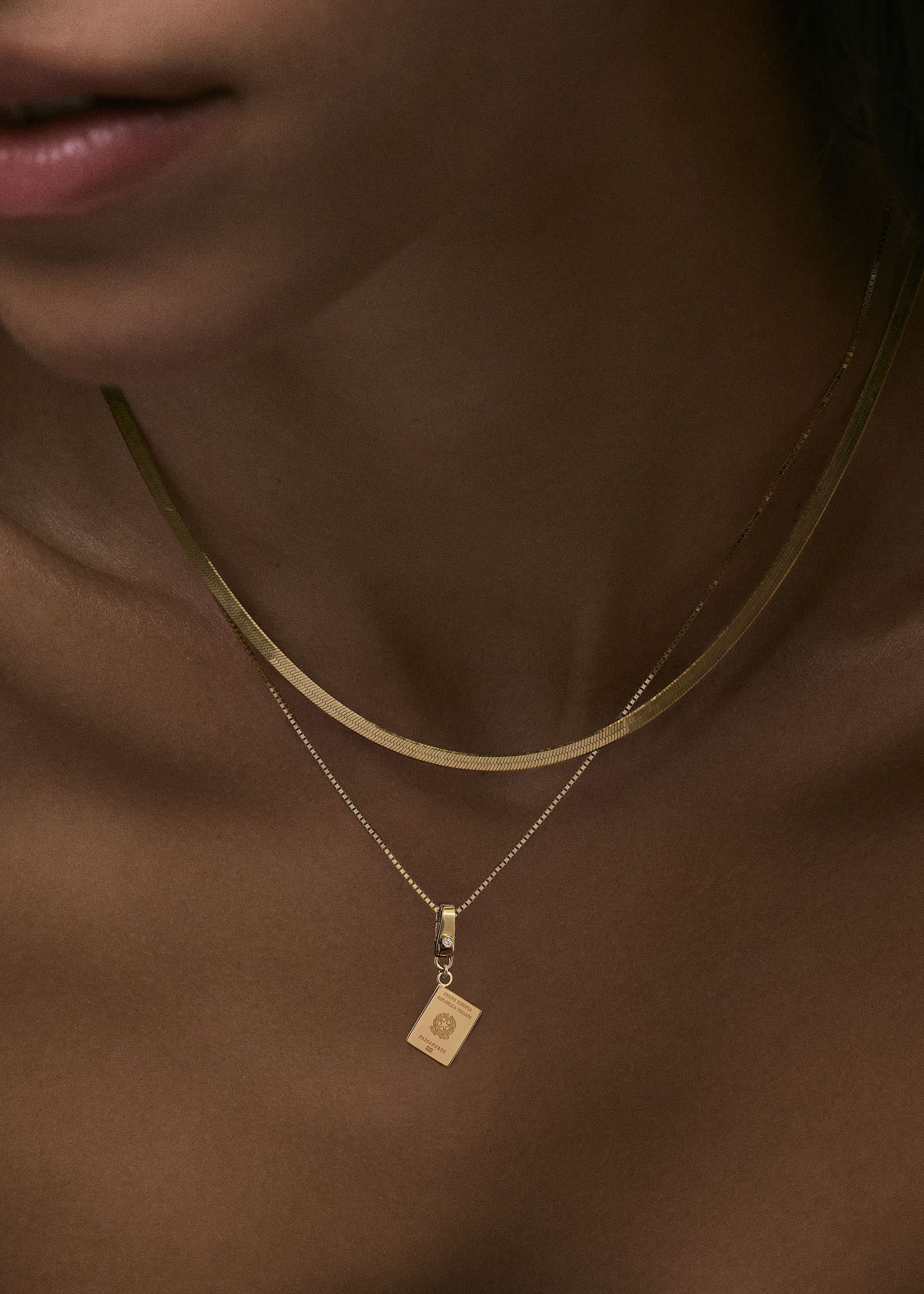 A close up of a Kinn 14k Gold Italian Heritage Passport Charm hanging off a Micro Rolo Chain Necklace layered with a Petite Carter Herringbone Chain Necklace