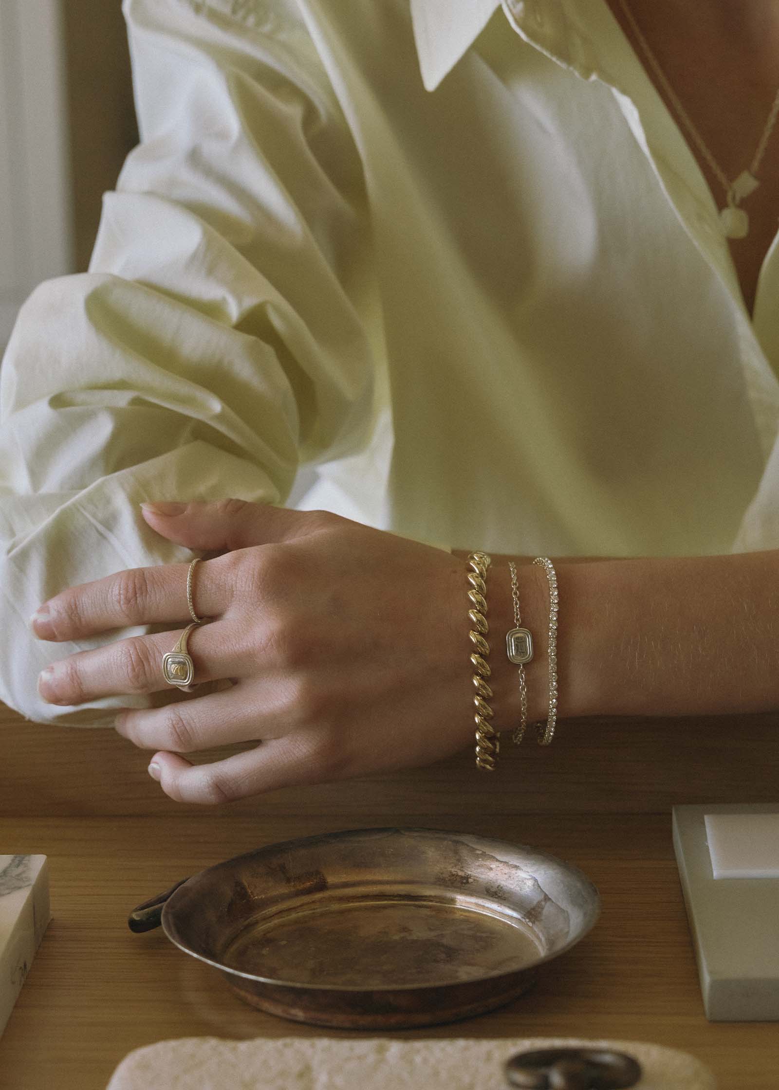 close up of a woman's hand over her desk wearing a Kinn x Shoppe Amber Interiors The Estate Bracelet Sterling Silver layered with a Diamond Tennis bracelet and Hampshire House bracelet along with The Estate Signet Ring on her middle finger